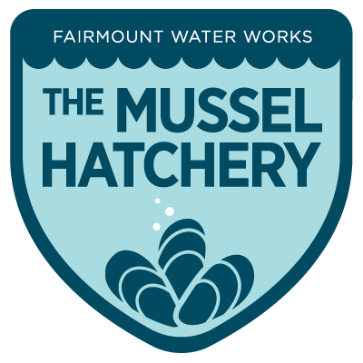 A shield-shaped badge with The Mussel Hatchery written large across the middle. The top says FAIRMOUNT WATER WORKS in small text, and there is a simple stylized illustration of three mussels at the bottom, resting in the point of the shield.