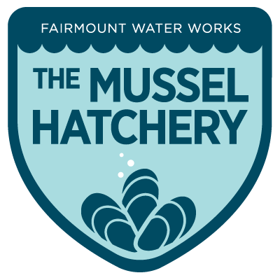 A shield-shaped badge with The Mussel Hatchery written large across the middle. The top says FAIRMOUNT WATER WORKS in small text, and there is a simple stylized illustration of three mussels at the bottom, resting in the point of the shield.