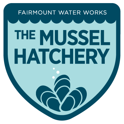 A shield-shaped badge with The Mussel Hatchery written large across the middle. The top says FAIRMOUNT WATER WORKS in small text, and there is a simple stylized illustration of three mussels at the bottom, resting in the point of the shield.