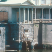 Historic photo of the Fairmount Water Works as seen from the river, with workers on a tall ladder and a board hanging on the face of the building over the water.