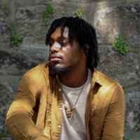 Jay Byrd, 2026 Alliance of Watershed Education Delaware River Watershed Fellow, wears a yellow shirt over a white t-shirt and sits looking to his right with one leg crossed over the other on a ledge in front of a stone wall with vines growing on it