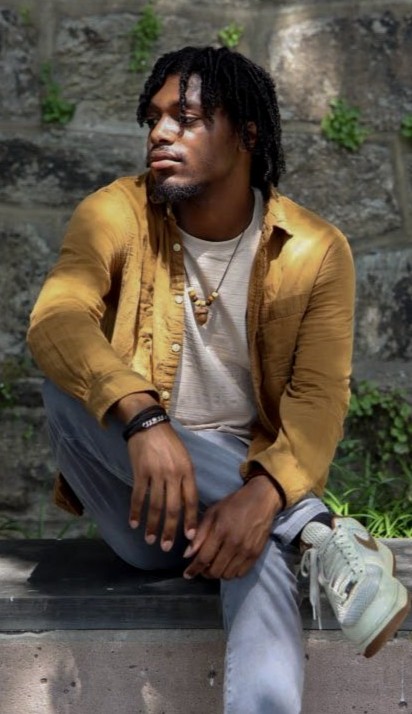 Jay Byrd, 2026 Alliance of Watershed Education Delaware River Watershed Fellow, wears a yellow shirt over a white t-shirt and sits looking to his right with one leg crossed over the other on a ledge in front of a stone wall with vines growing on it