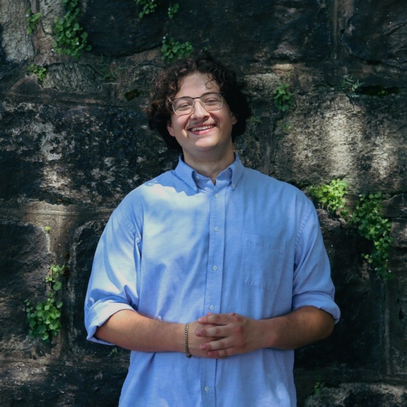 Michael De La Parra Gurr, 2026 Interpretive Center Summer Intern, wears a blue shirt and smiles as he stands in front of a shaded stone wall with vines growing on it
