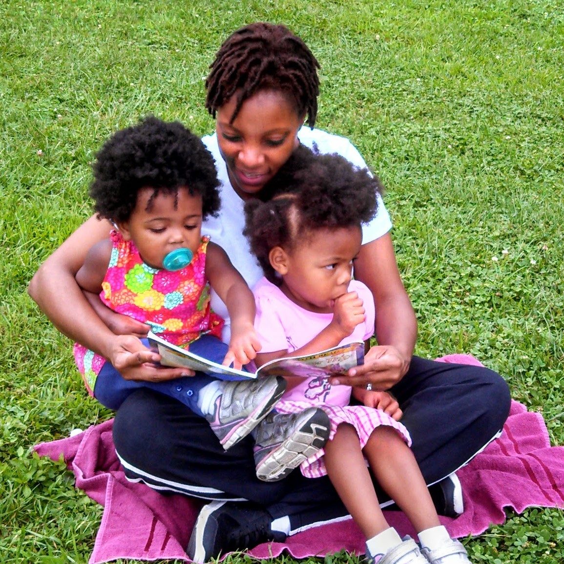 photo of a woman sitting in the grass reading to the two little girls in her lap